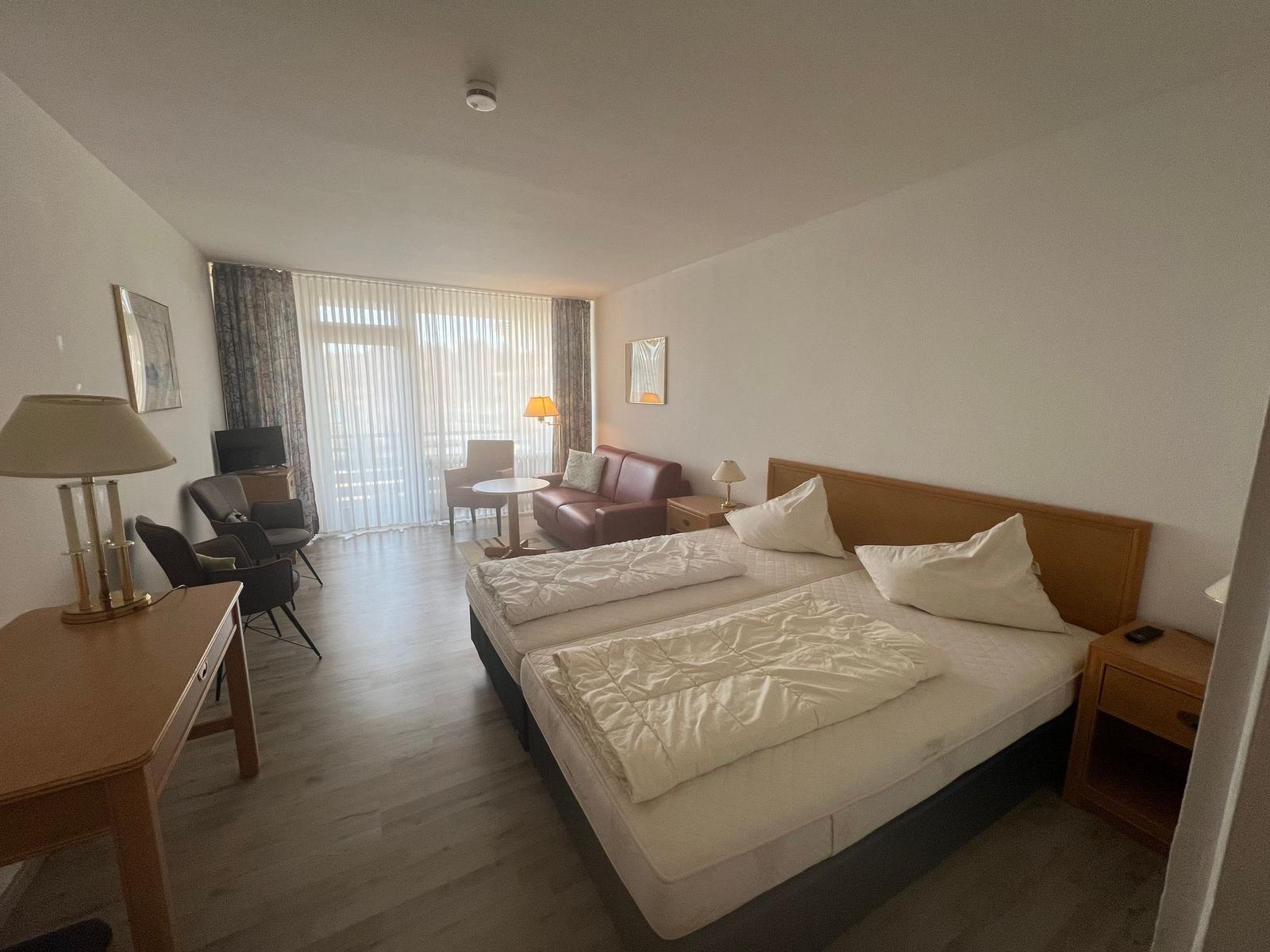 Zimmer mit Doppelbett, Sofa, Schreibtisch und TV. Blick auf Balkon durch Fenster.