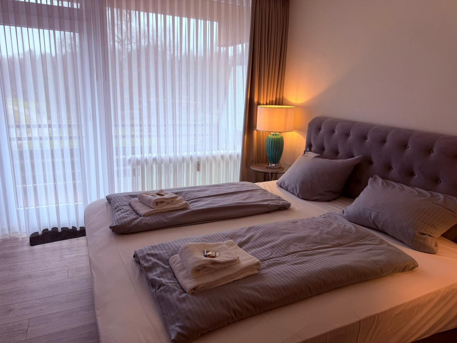 Doppelbett mit Bettwäsche, Nachttisch und Lampe neben großem Fenster mit Jalousie.