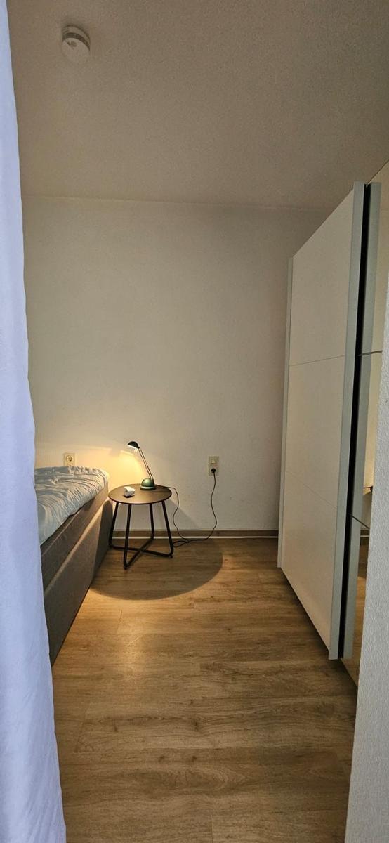 Schlafzimmer mit Bett, Nachttisch und Lampe. Holzboden und Schrank sichtbar.
