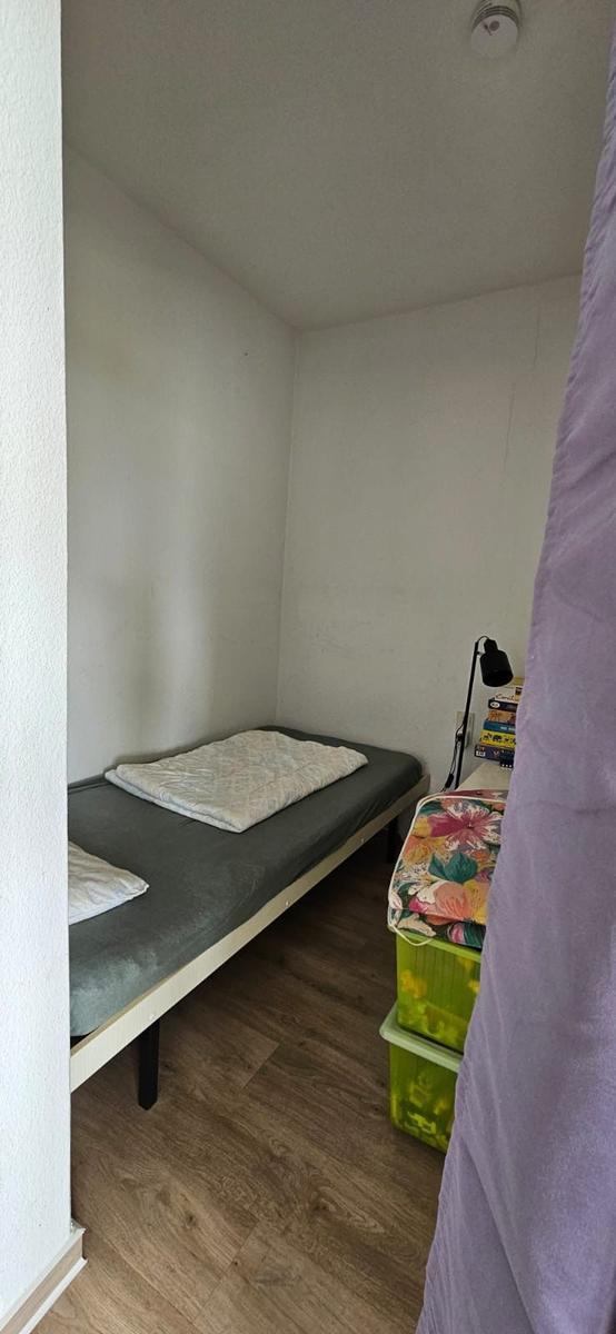 Schlafzimmer mit Bett, Nachttisch und Lampe. Holzfußboden und weiße Wände.