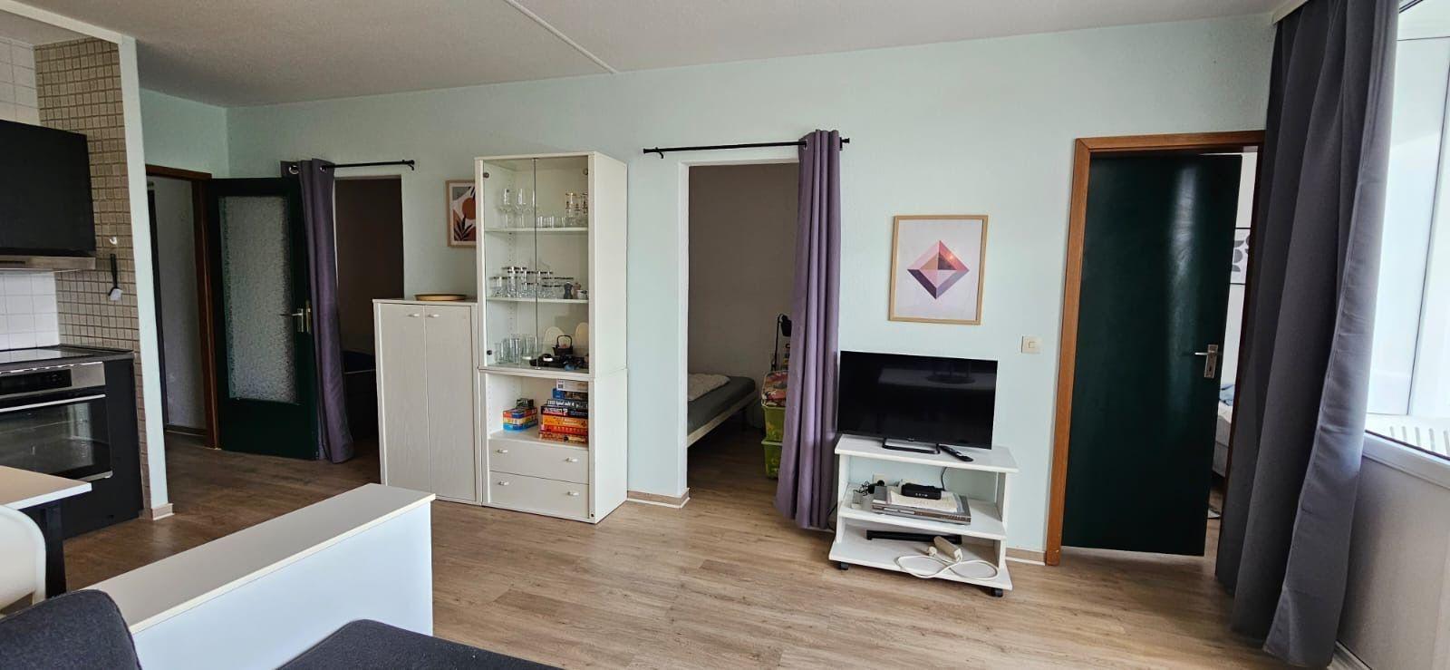 Wohnung mit Küche, Wohnbereich, Fernseher und Schlafzimmer. Holzboden und weiße Möbel.