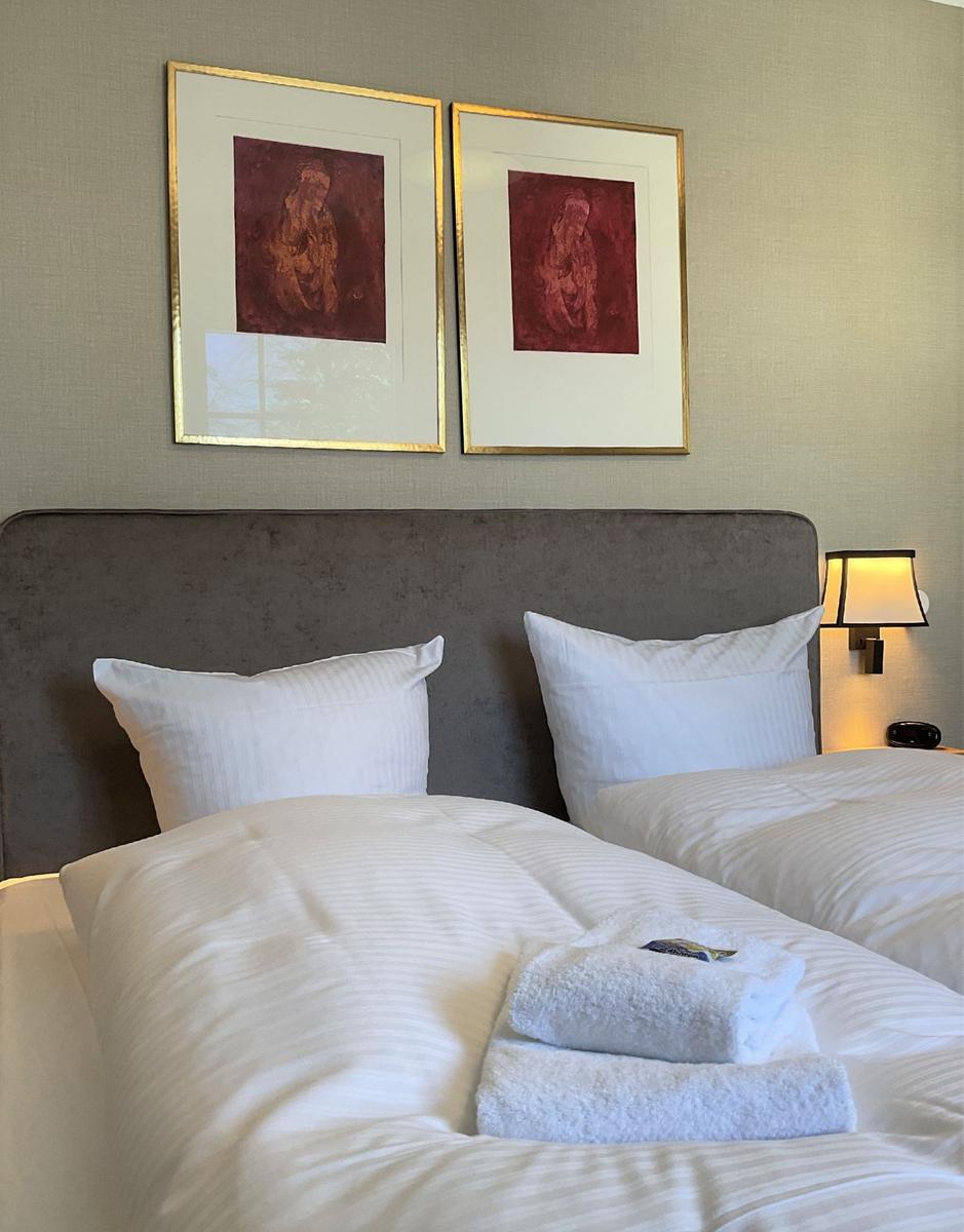 Ein Bett mit weißen Laken und zwei Kissen. Über dem Bett hängen zwei Bilder in goldenen Rahmen.