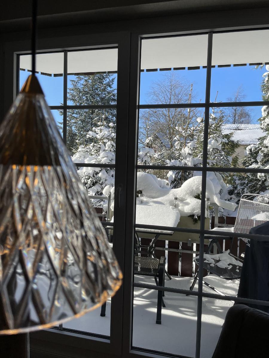 Durch die Glastür blickt man auf eine verschneite Terrasse mit Gartenmöbeln und schneebedeckten Bäumen.