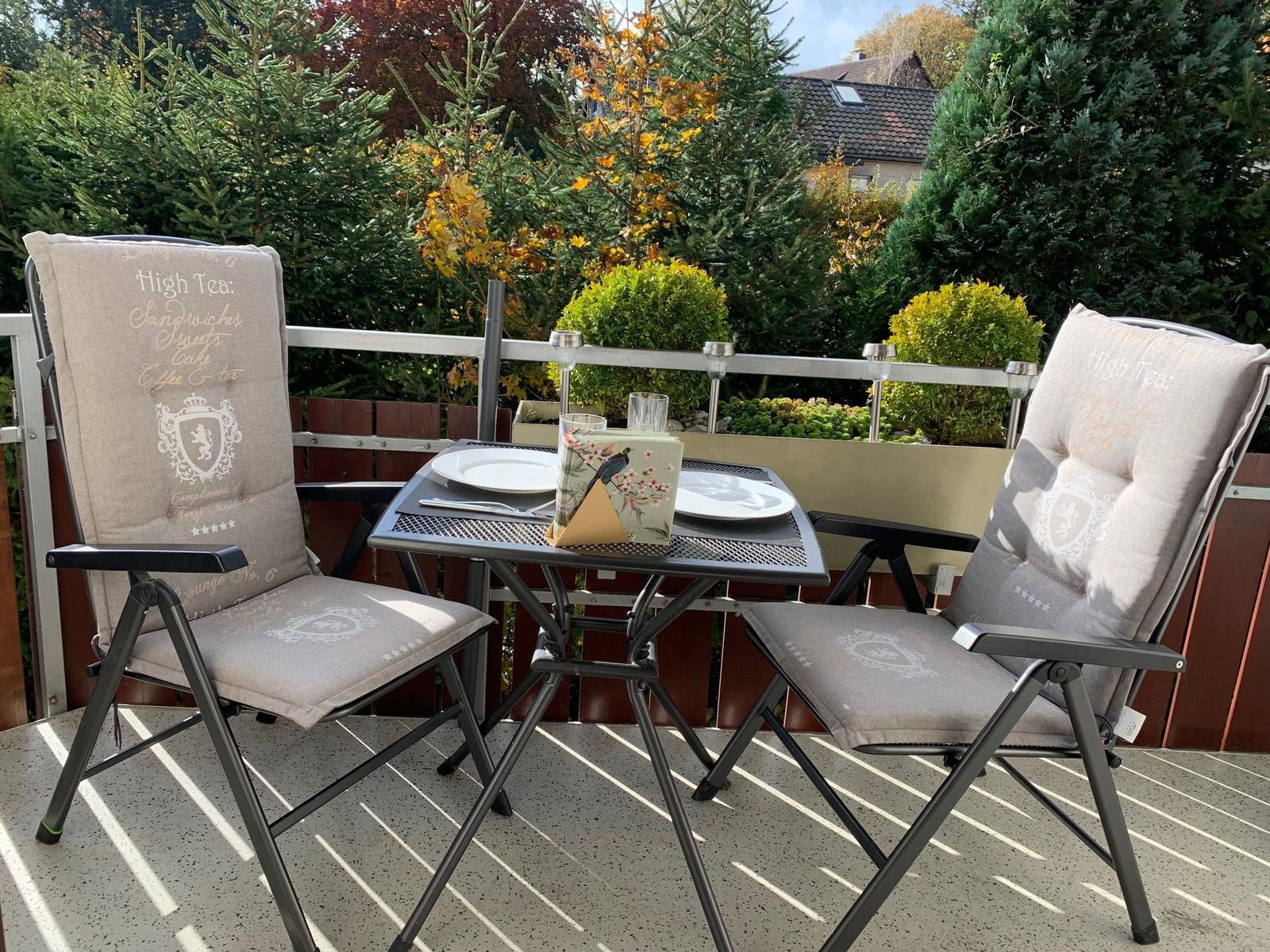 Terrasse mit Tisch und zwei Stühlen, umgeben von Bäumen und Sträuchern.