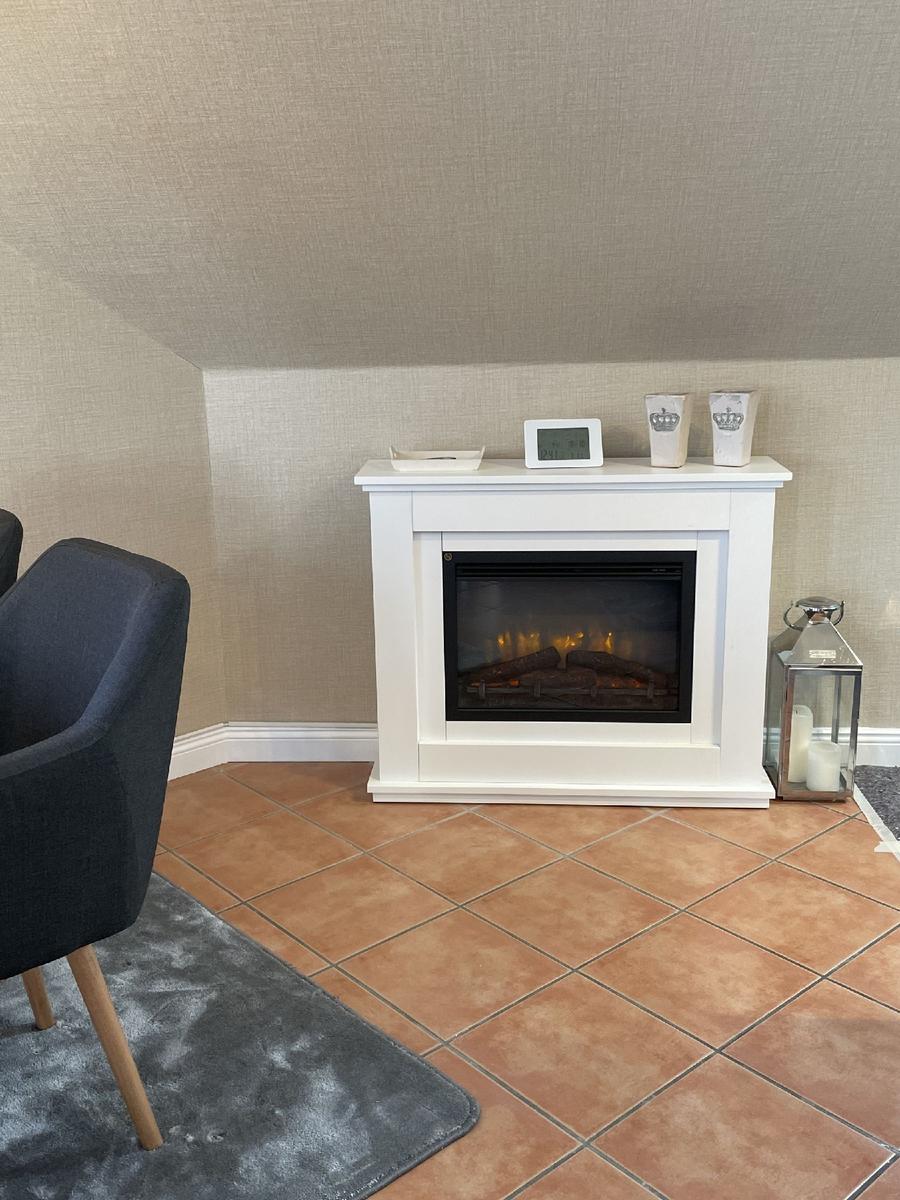 Weißer Kamin mit brennendem Kaminofen, daneben Stehlampe und Sitzgelegenheit.