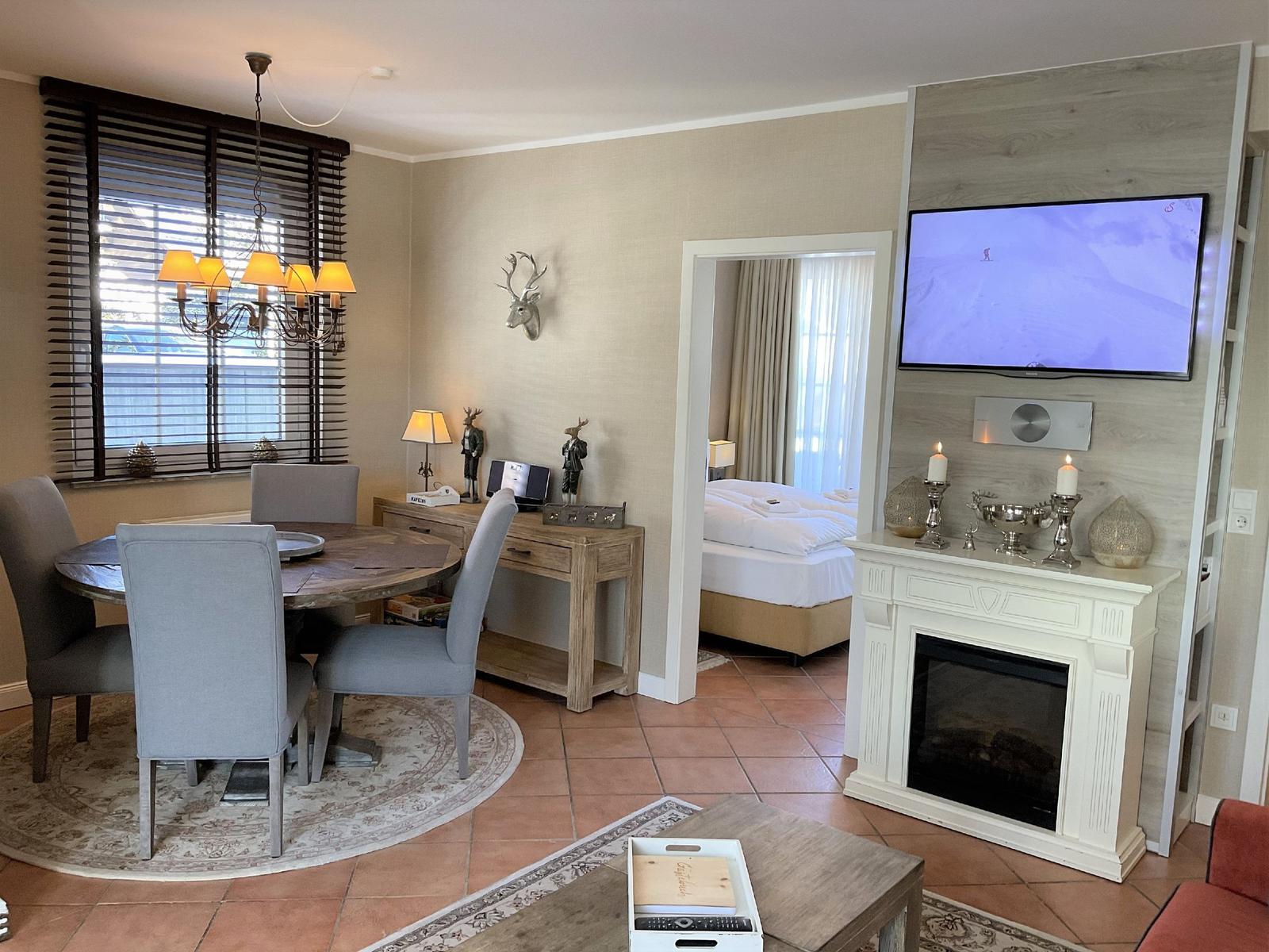 Wohn- und Essbereich mit Schlafzimmer im Hintergrund. Fernseher und Kamin im Vordergrund.