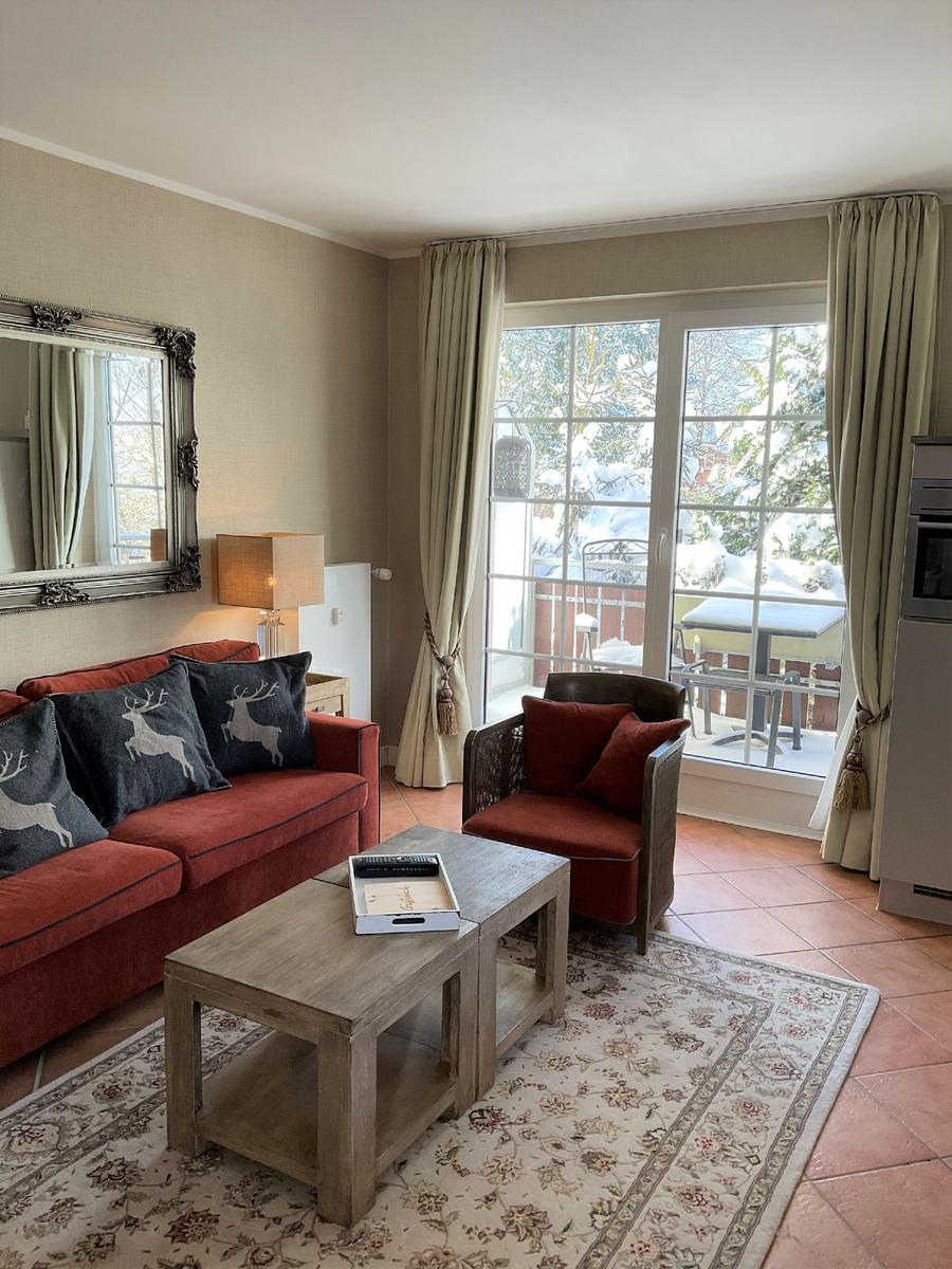 Wohnzimmer mit Sofa, Sessel und Terrasse. Durchgehende Glasfront mit Blick auf verschneite Bäume.