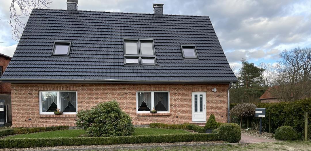 Ein zweistöckiges Haus mit rotem Backstein und dunklem Dach. Vorgarten mit Beeten und Sträuchern.