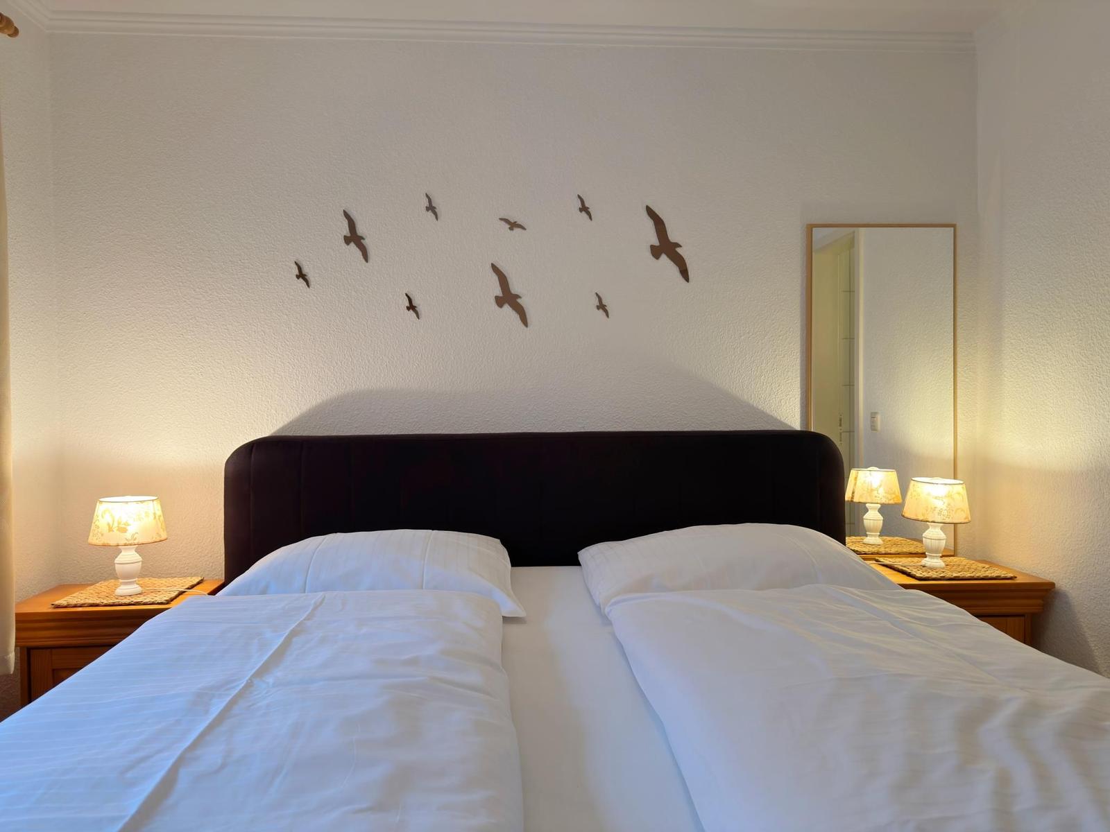 Doppelbett mit weißen Bettwäsche, Nachttische mit Lampen und Wanddekoration aus Vögeln.