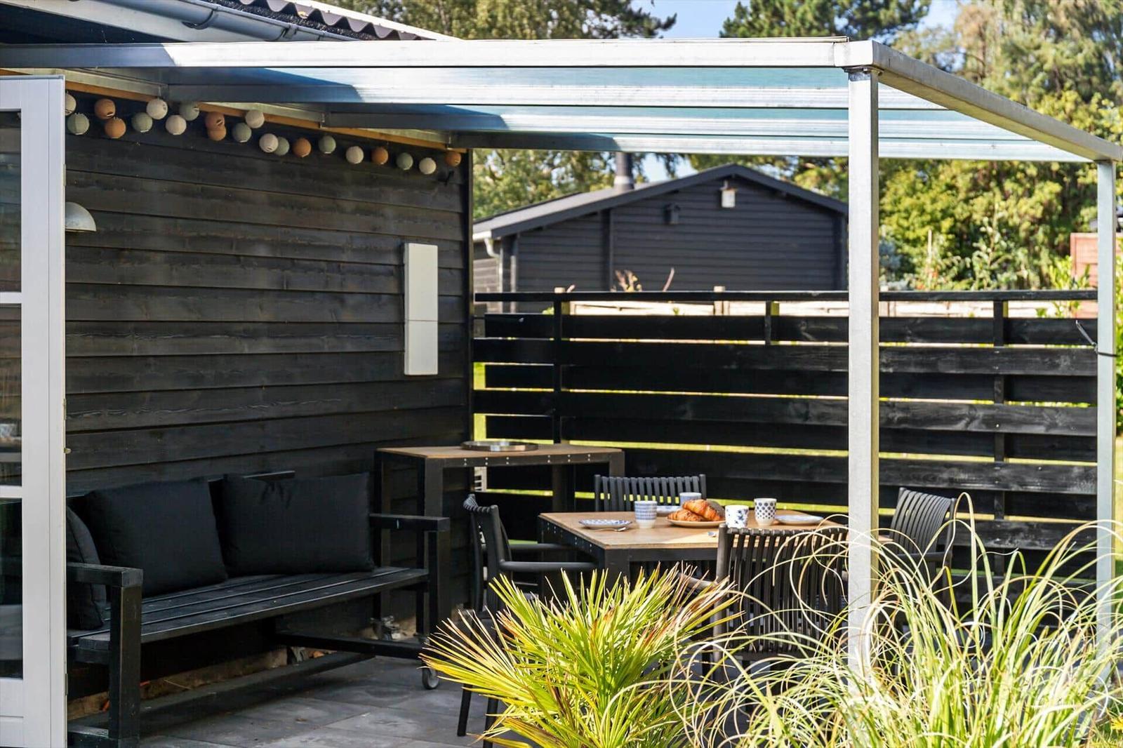 Terrace with seating and table under a canopy. Black wooden wall and plants in the foreground.