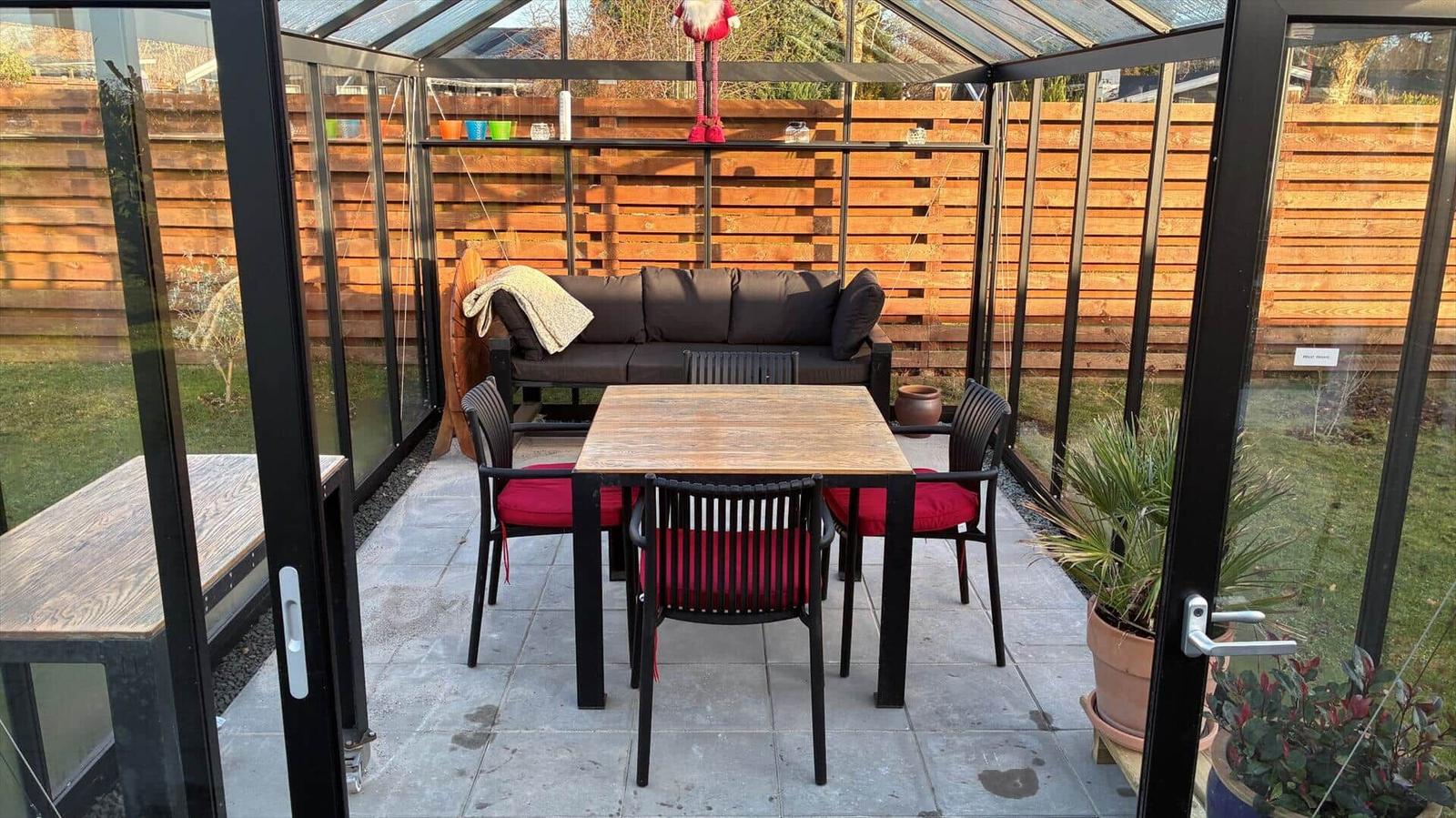 Glass conservatory with seating group, table and chairs. Wooden table, red cushions, plants.