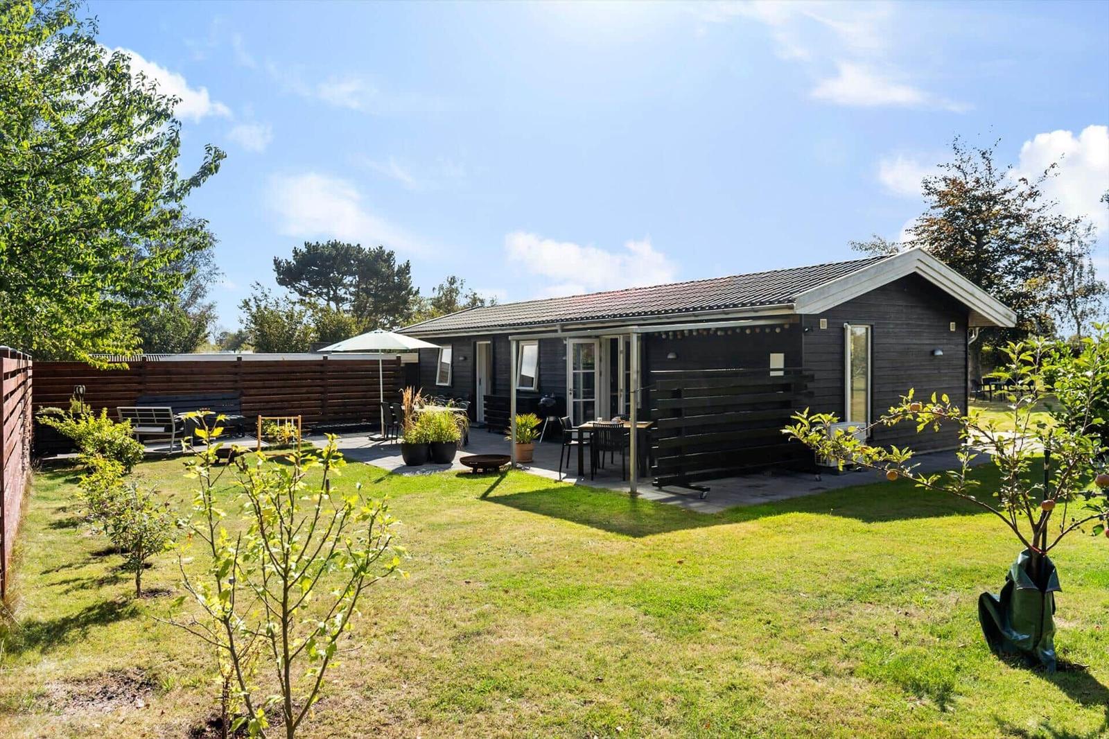 A dark wooden house with terrace, garden, and barbecue area under blue sky.