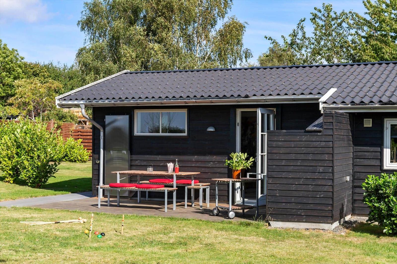 Black wooden house with terrace, table, benches, and grill area in the garden.