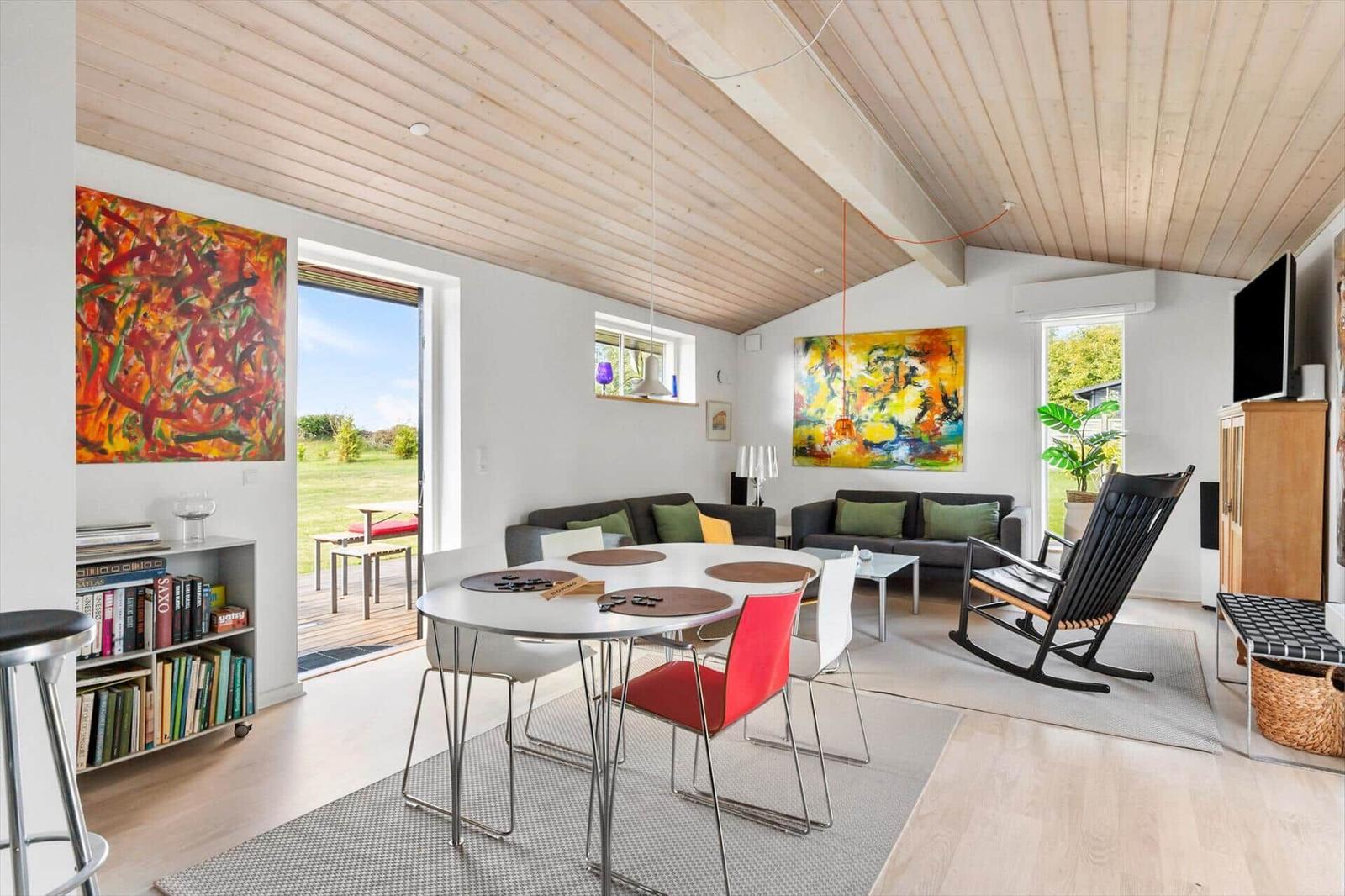 Living and dining area with table, chairs, sofa, and wall art. Wooden ceiling and large windows.