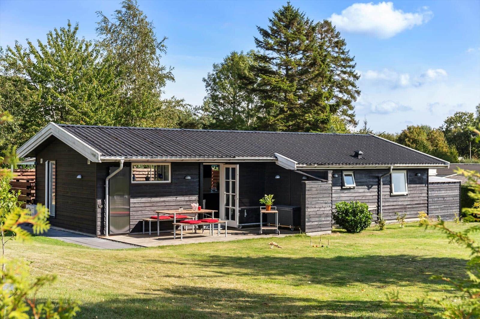 Wooden house with terrace, grill, and garden. Table with red cushions. Surrounded by trees.