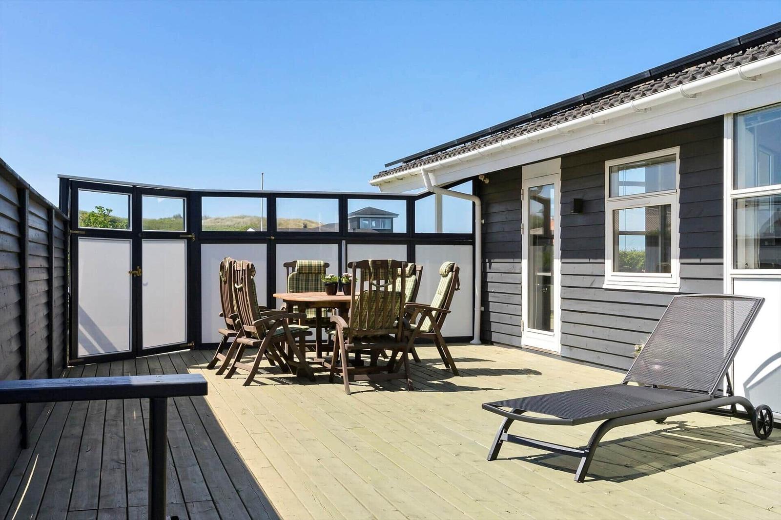 Deck with wooden table, chairs, and lounge chair. Background: house with windows and view of landscape.