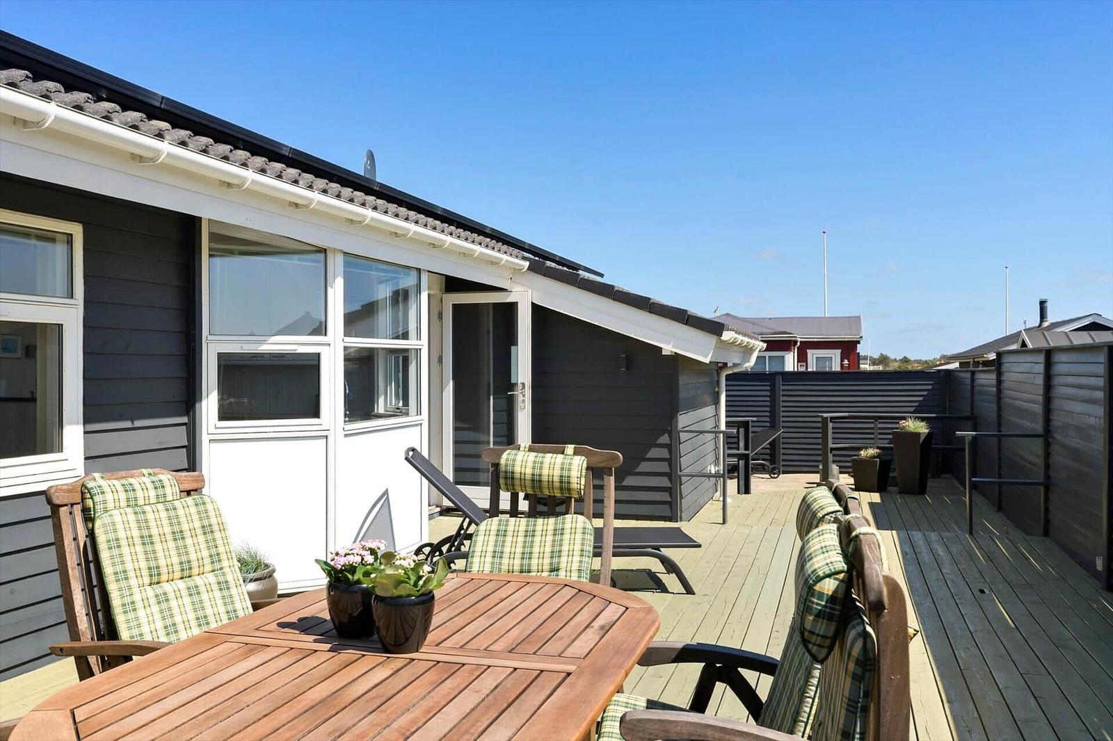 Deck with wooden table, chairs, and sun loungers. Background: house with windows and fence.