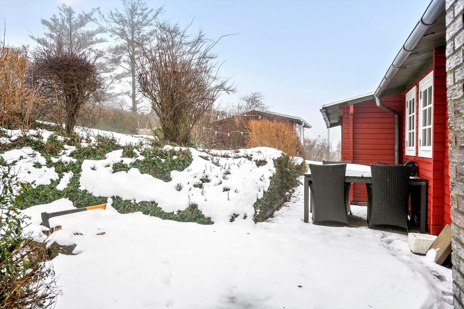 Roter Holzhaus mit Tisch und Stühlen im Schnee. Garten mit Sträuchern und Bäumen.