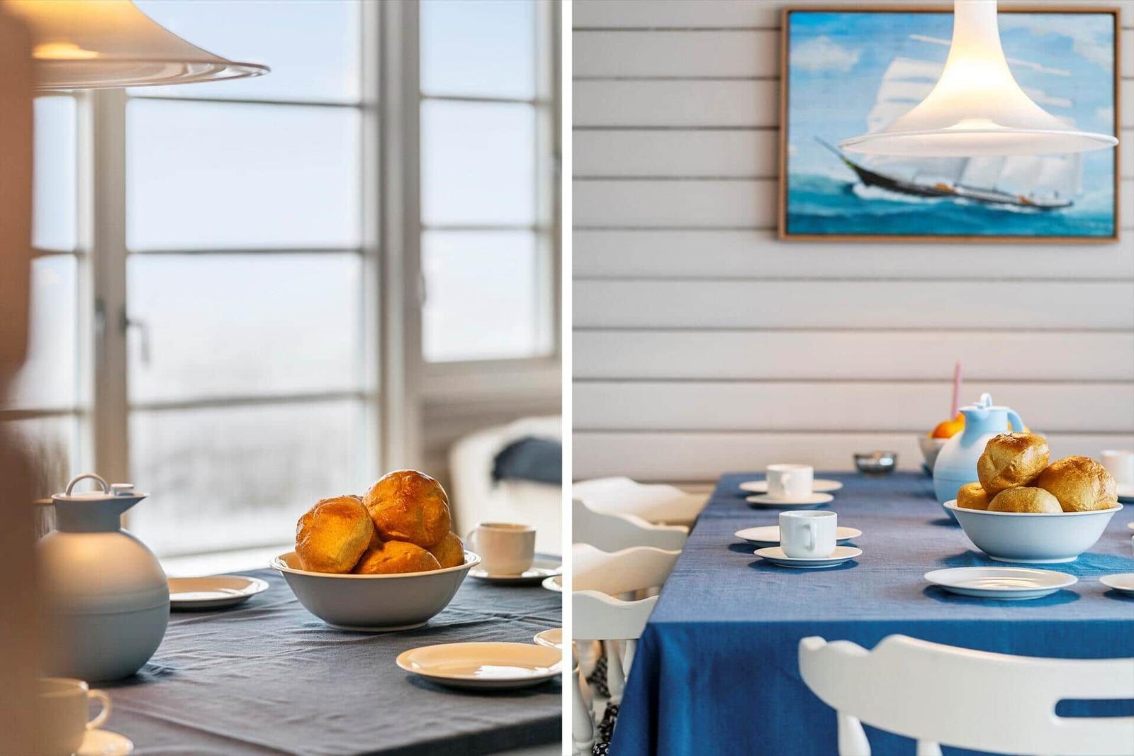 Esstisch mit blauer Tischdecke, Kaffeetassen und Brötchen. Wandbild mit Segelschiff.