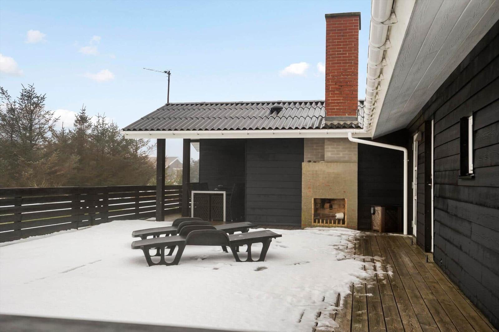 Terrasse mit Schnee, Sitzgelegenheiten und Holzdecke. Im Hintergrund steht ein kleines Gebäude mit Schornstein.