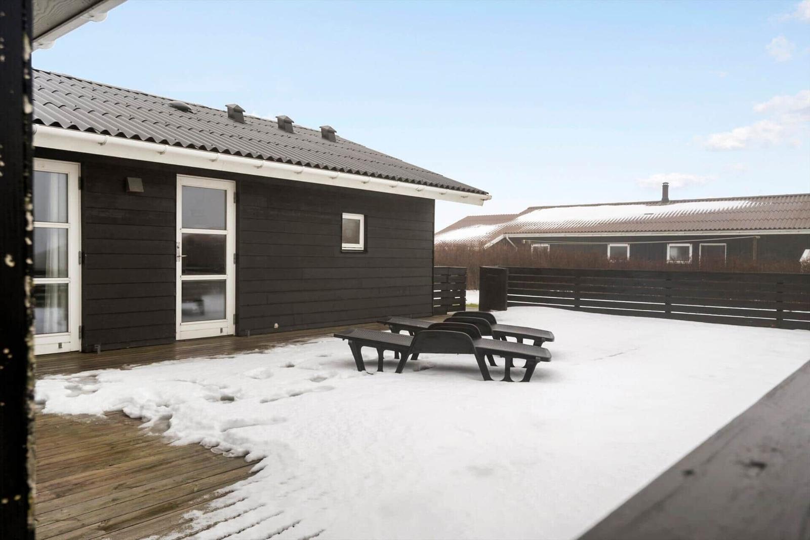 Schneebedeckter Außenbereich mit Liegen vor einem dunklen Holzhaus mit weißer Tür.