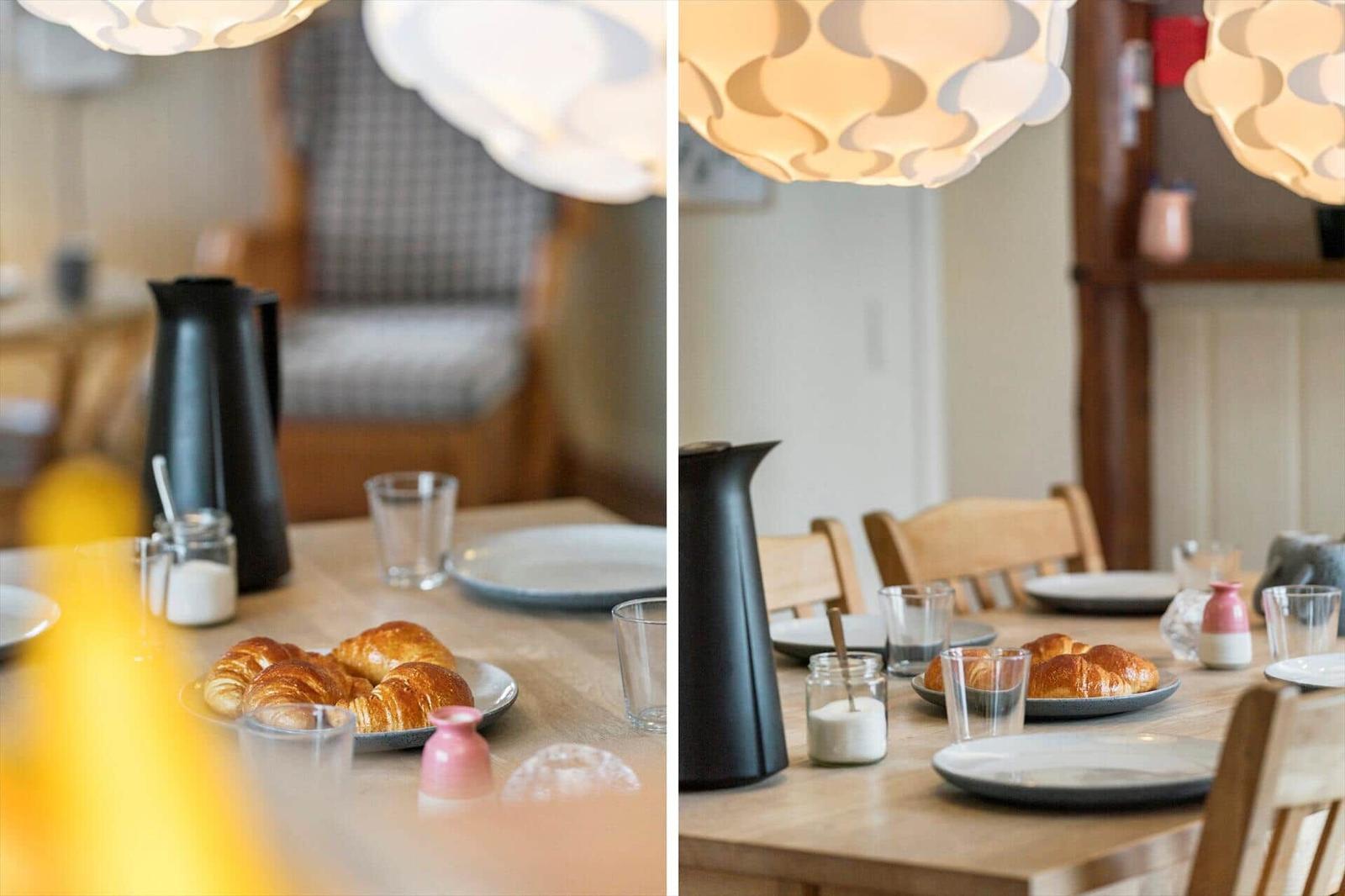 Esstisch mit Croissants, Gläsern und einer schwarzen Kanne unter einer Lampe.