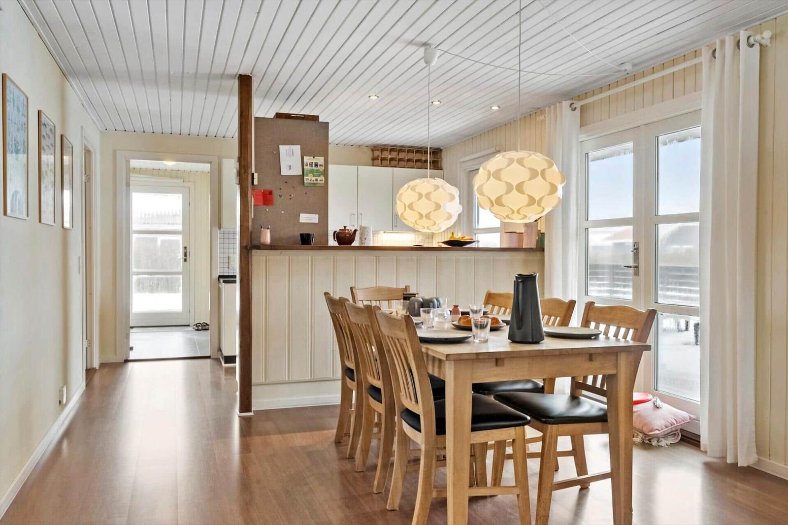 Essbereich mit Holztisch, Stühlen und zwei hängenden Lampen. Küche im Hintergrund. Holzfußboden und Fenster.