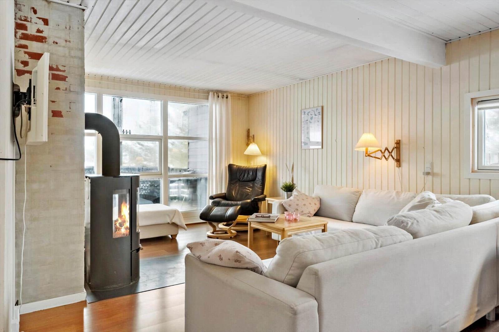 Wohnzimmer mit Sofa, Sessel und Kaminofen. Holzwände und große Fenster.