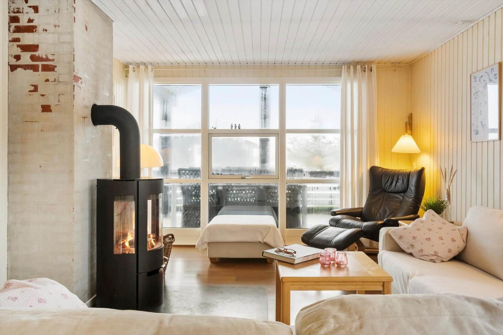 Wohnraum mit Kamin, Sofa, Sessel und großem Fenster. Holzfußboden und hell gestrichene Wände.