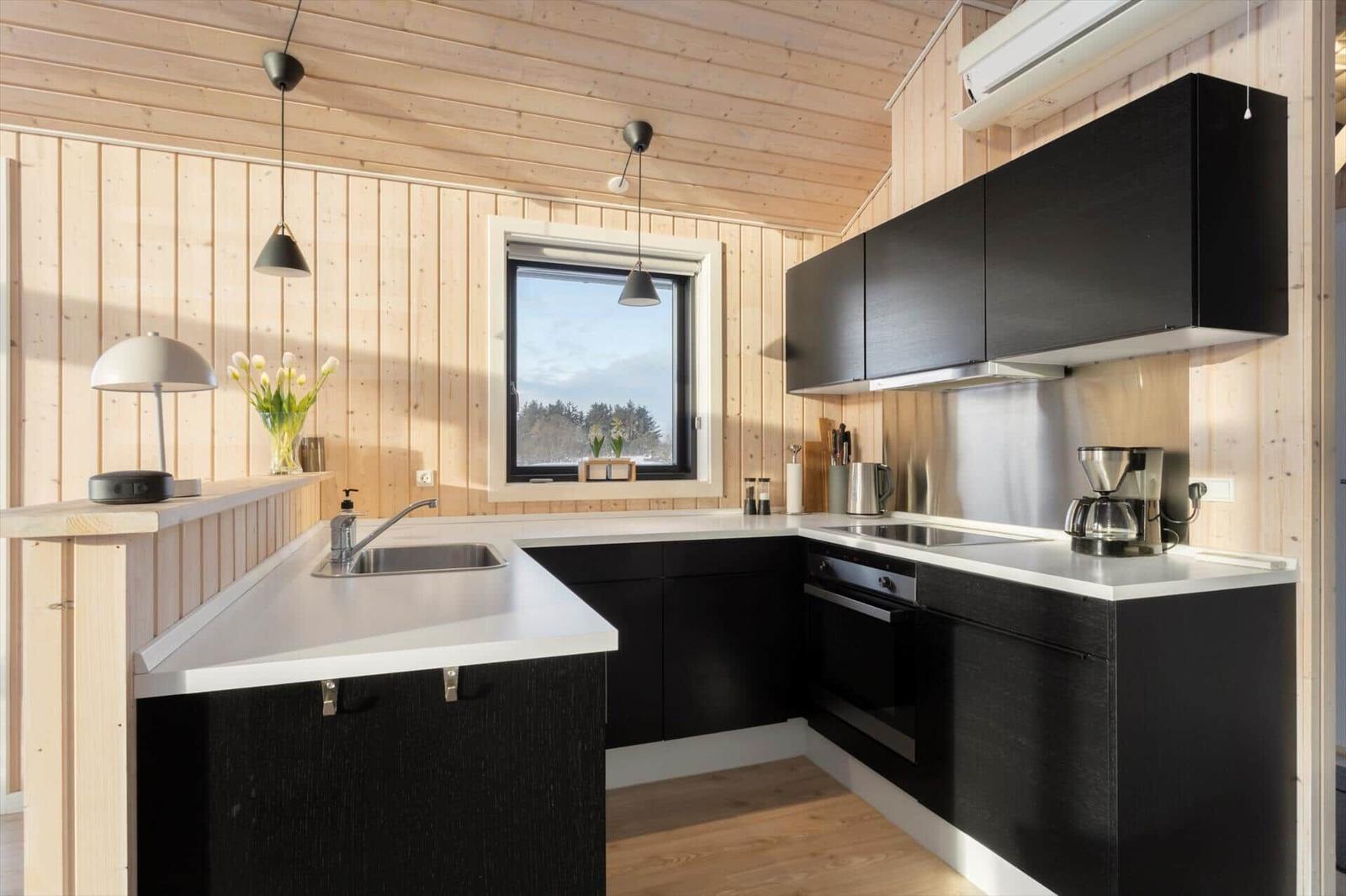 Kitchen with black cabinets, white countertops, and wood-paneled walls. Window with view of forest.