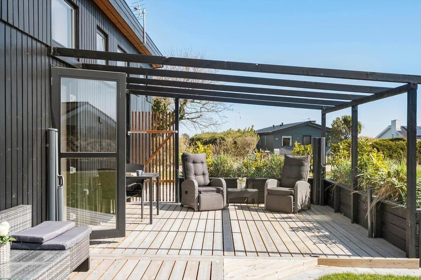 Terrasse mit Pavillon, zwei Sessel und Tisch. Holzdecke und Pflanzen umgeben den Außenbereich.