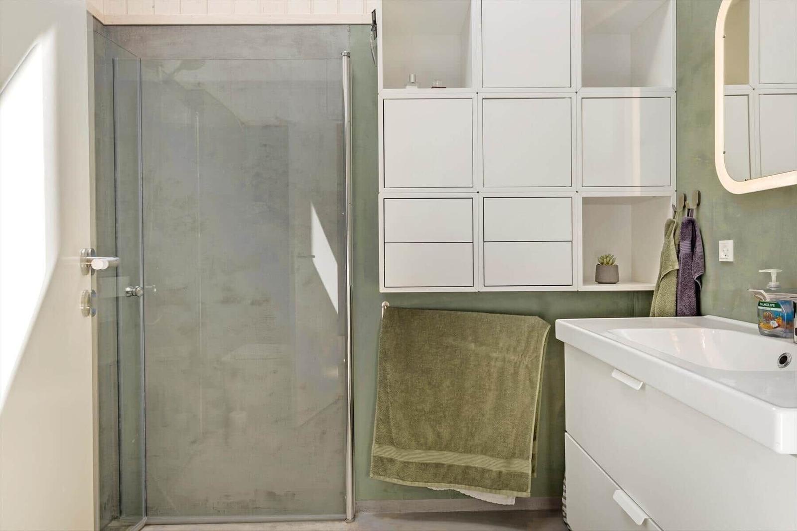 Badezimmer mit Dusche, Waschbecken und weißen Schränken an grünem Wandpaneel.