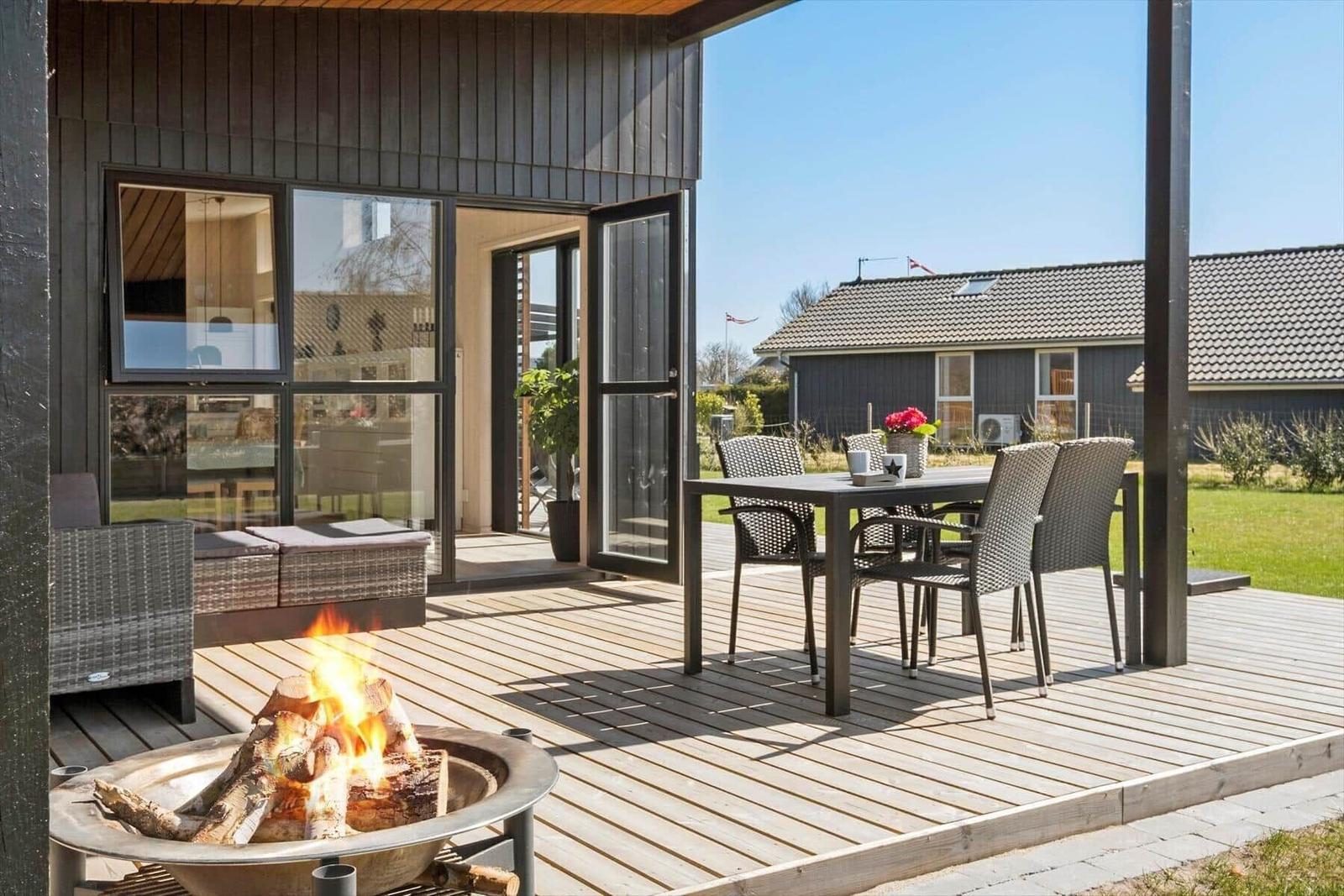 Terrasse mit Sitzgruppe, Esstisch und brennendem Feuerkorb. Hintergrund: Garten und Nachbarhaus.