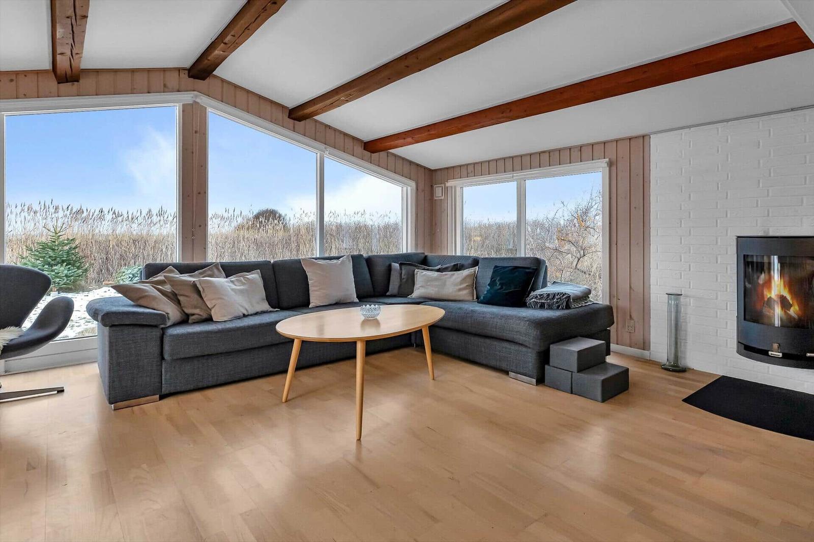 Wohnzimmer mit Sofa, Kamin und großen Fenstern. Holzboden und Balken an der Decke.
