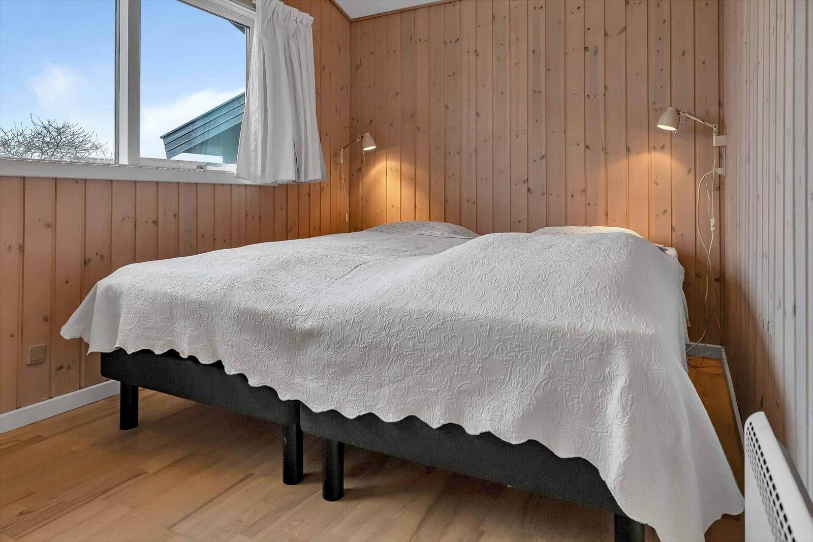Schlafzimmer mit Bett, Holzwand und Fenster. Zwei Wandlampen und Heizkörper sind sichtbar.