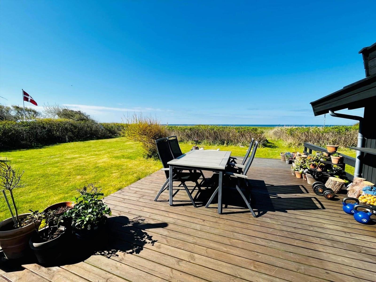 Terrasse mit Tisch und Stühlen, Blick auf Grünland und Meer, daneben Flagge und Pflanzen.