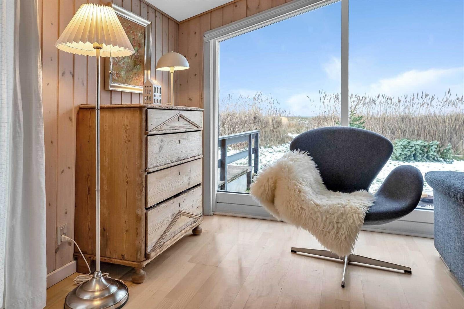 Ein Raum mit Holzwand, Schrank, Stehlampe und Sessel vor großem Fenster mit Aussicht.