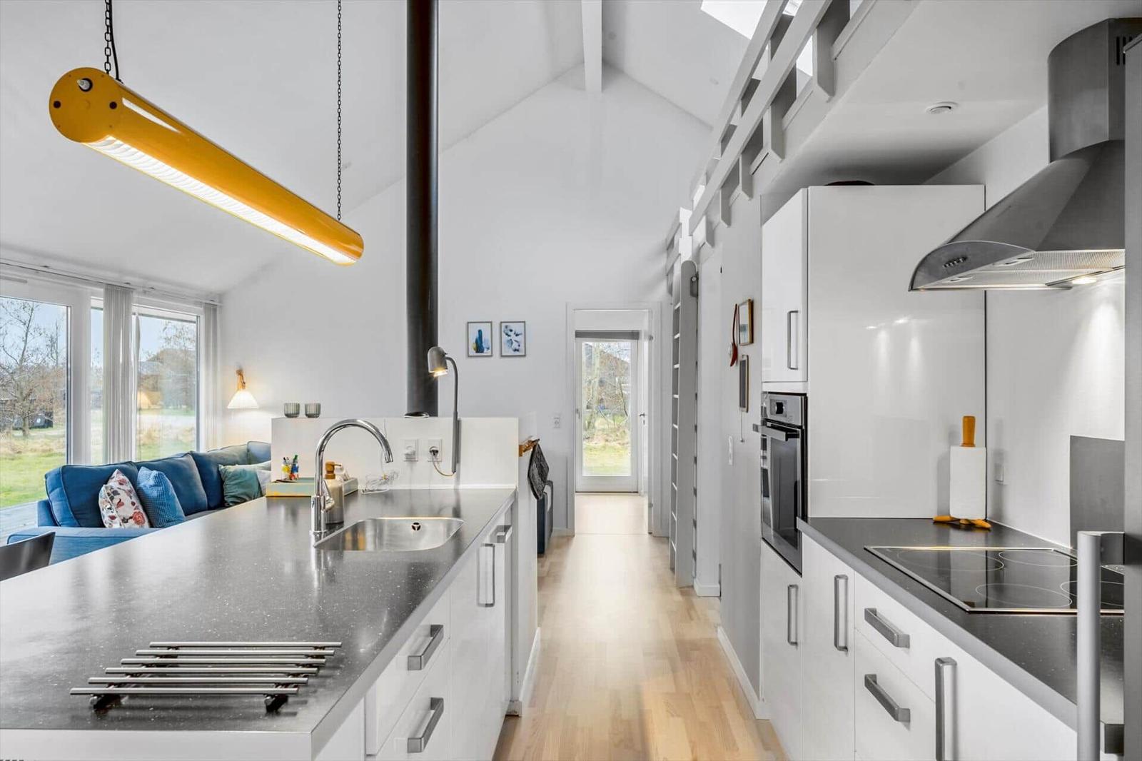 Modern kitchen with countertop, sink, and cooktop. Living area with sofa and windows.
