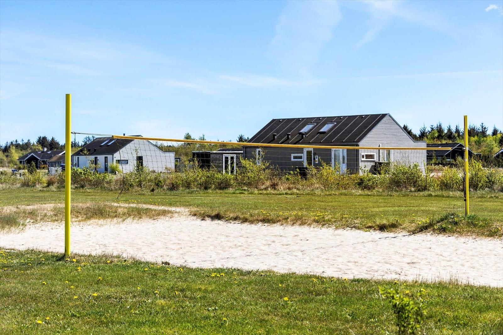 Volleyball net in front of beach house with green field and sand. Background: holiday homes and trees.