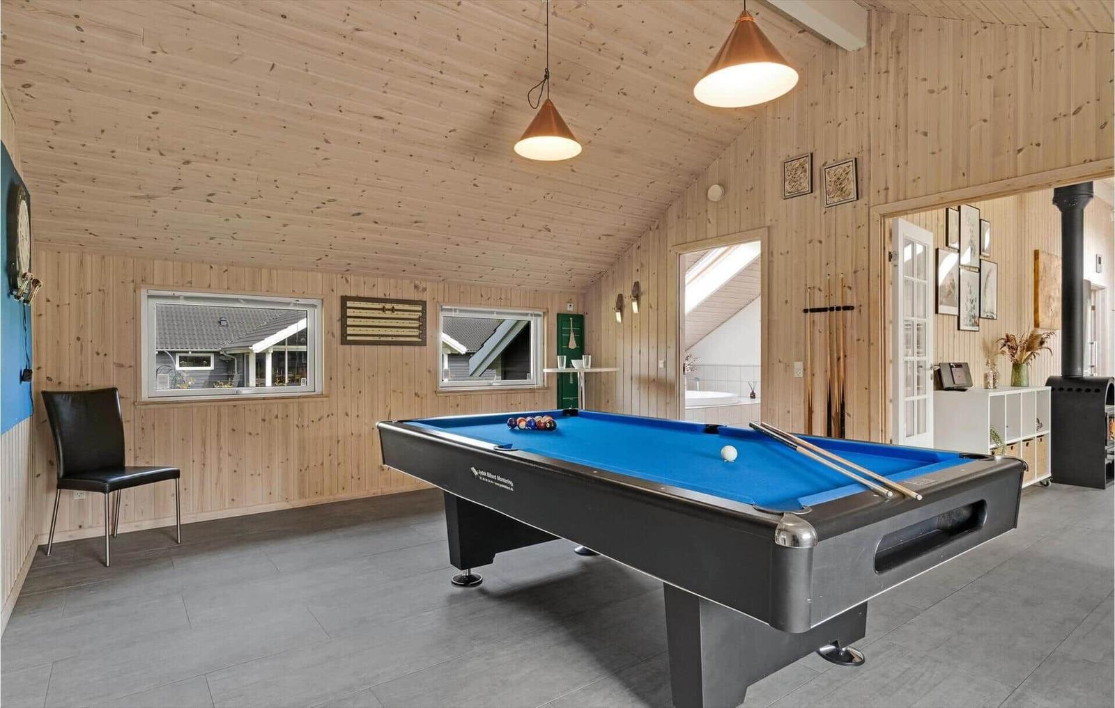 Room with pool table, wooden walls and windows. Bathroom and fireplace in background.