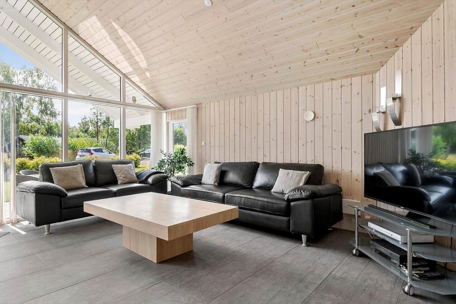 Living room with two black sofas, wooden table, and TV. Wooden walls and large windows.