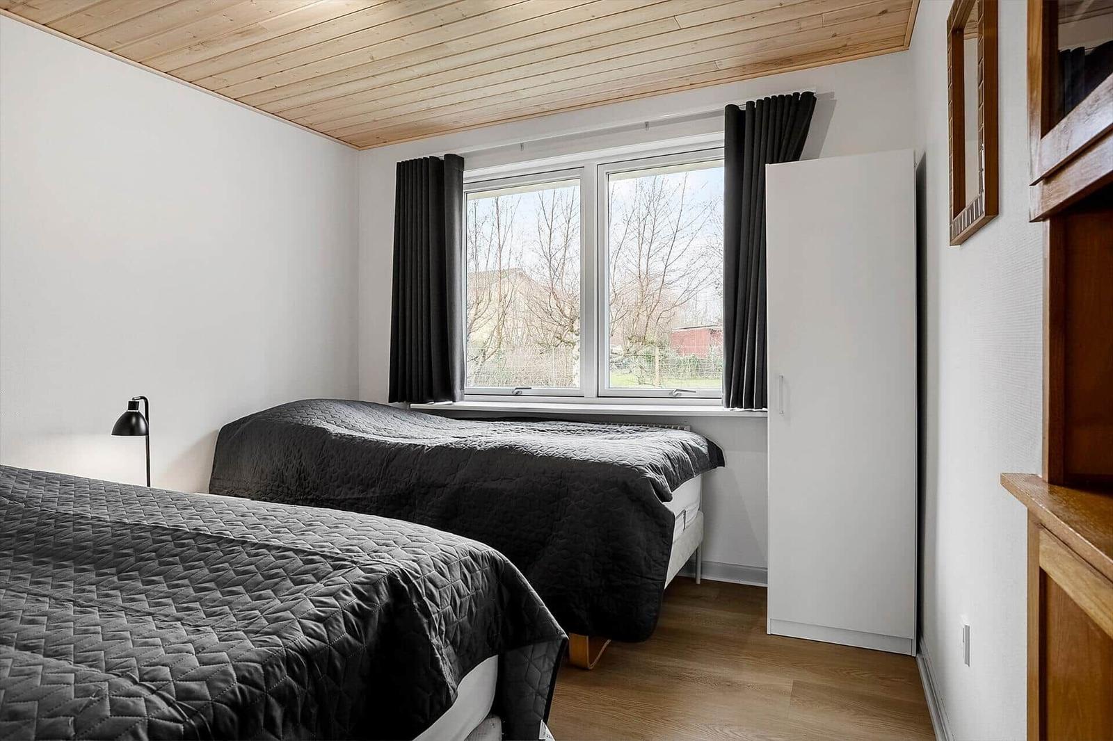 Zwei Betten mit dunklen Bezügen, Fenster mit schwarzen Vorhängen und Holzdecke.