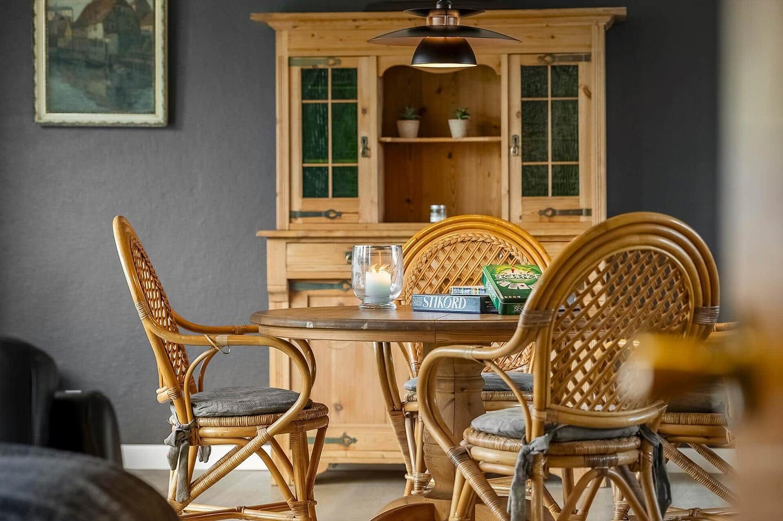 Rattan-Stuhl um einen Holztisch mit Kerze und Spielboxen. Hintergrund: Holzschrank und Wandbild.