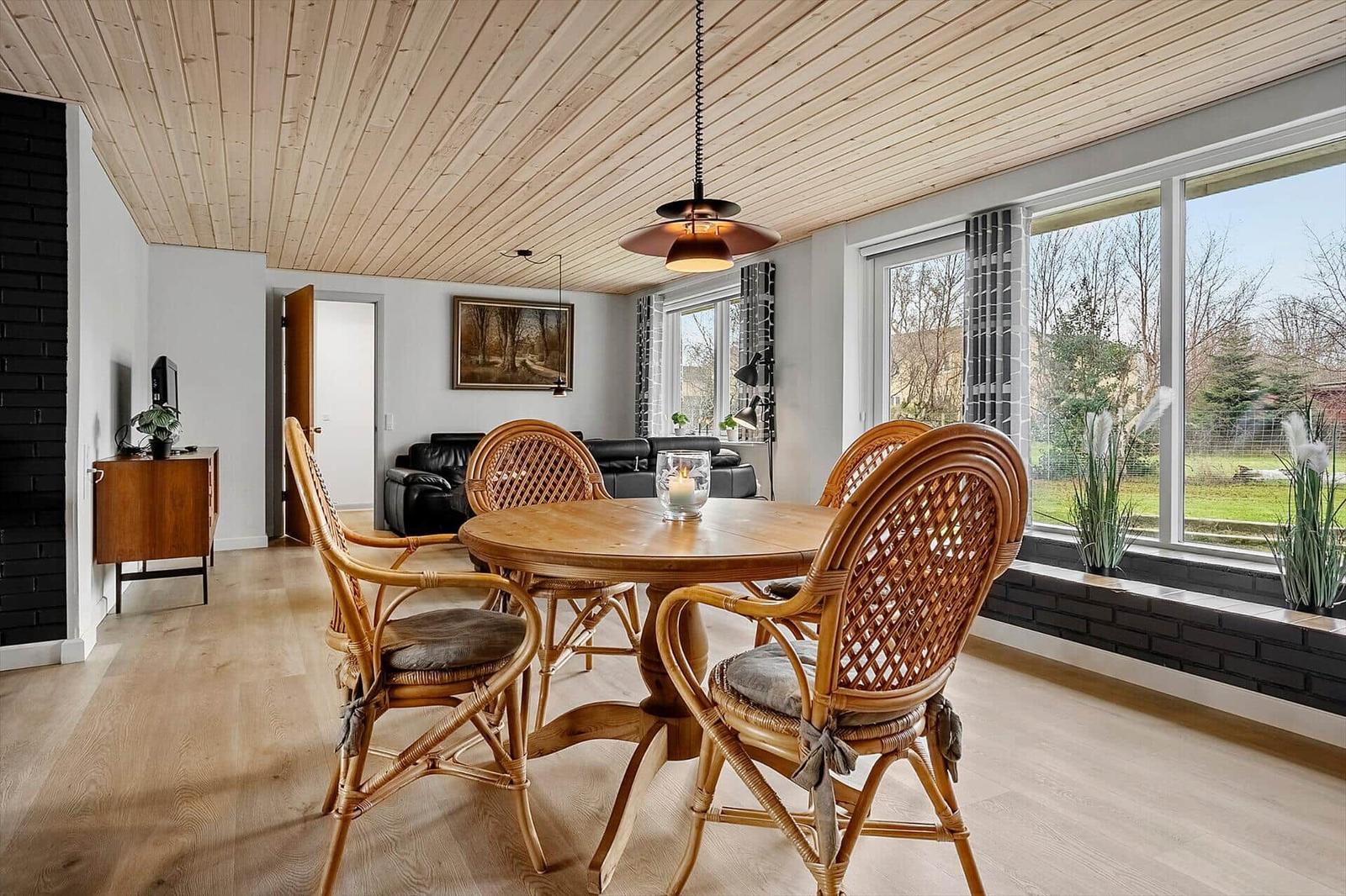 Esszimmer mit Rattan-Stühlen und Holztisch. Große Fenster und Holzdecke.