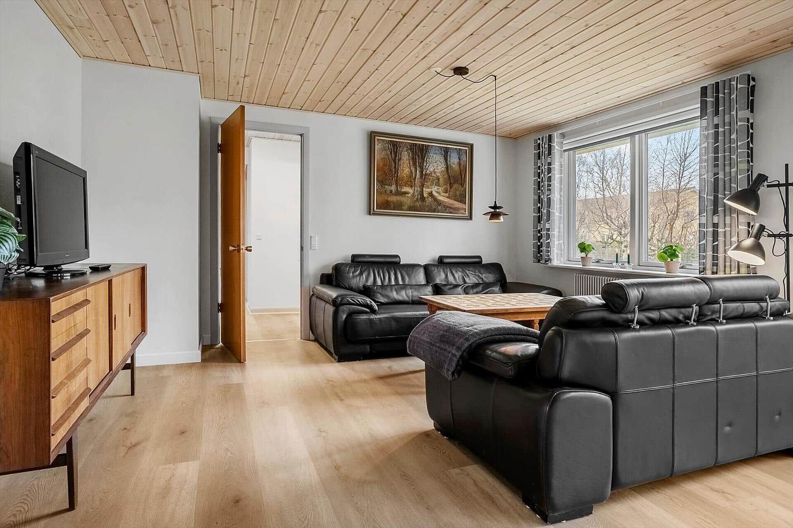 Wohnzimmer mit zwei schwarzen Ledersofas, Holzboden und Holzdecke. Fenster mit Vorhängen und Fernseher.