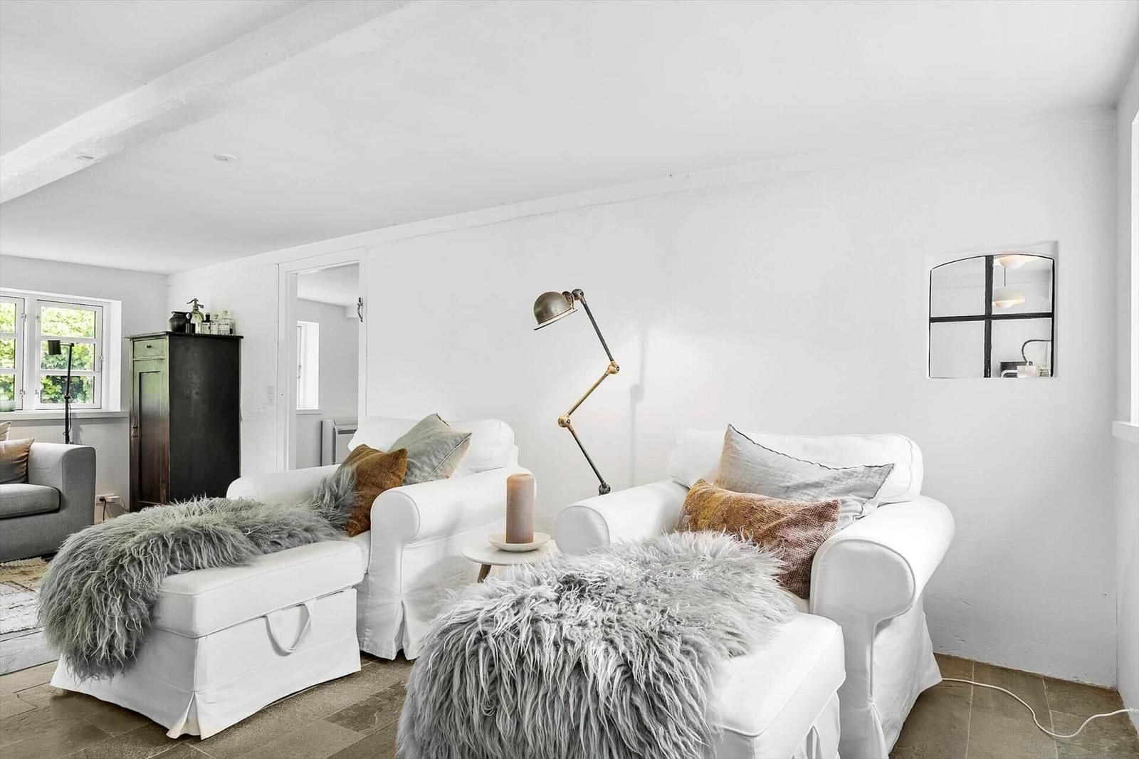 Wohnzimmer mit weißen Sesseln, Felldecken und Stehlampe. Fenster und Tür sind sichtbar.