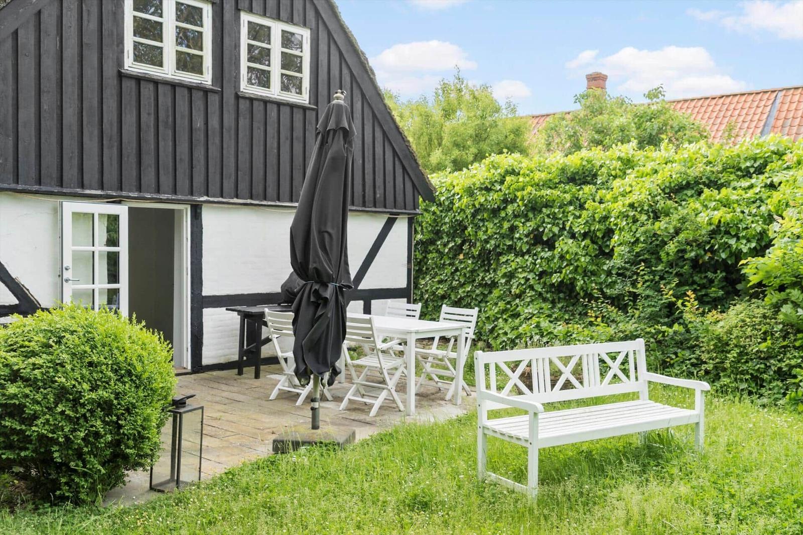 Terrasse mit Tisch, Stühlen und Bank vor schwarz-weißem Haus mit grüner Hecke.