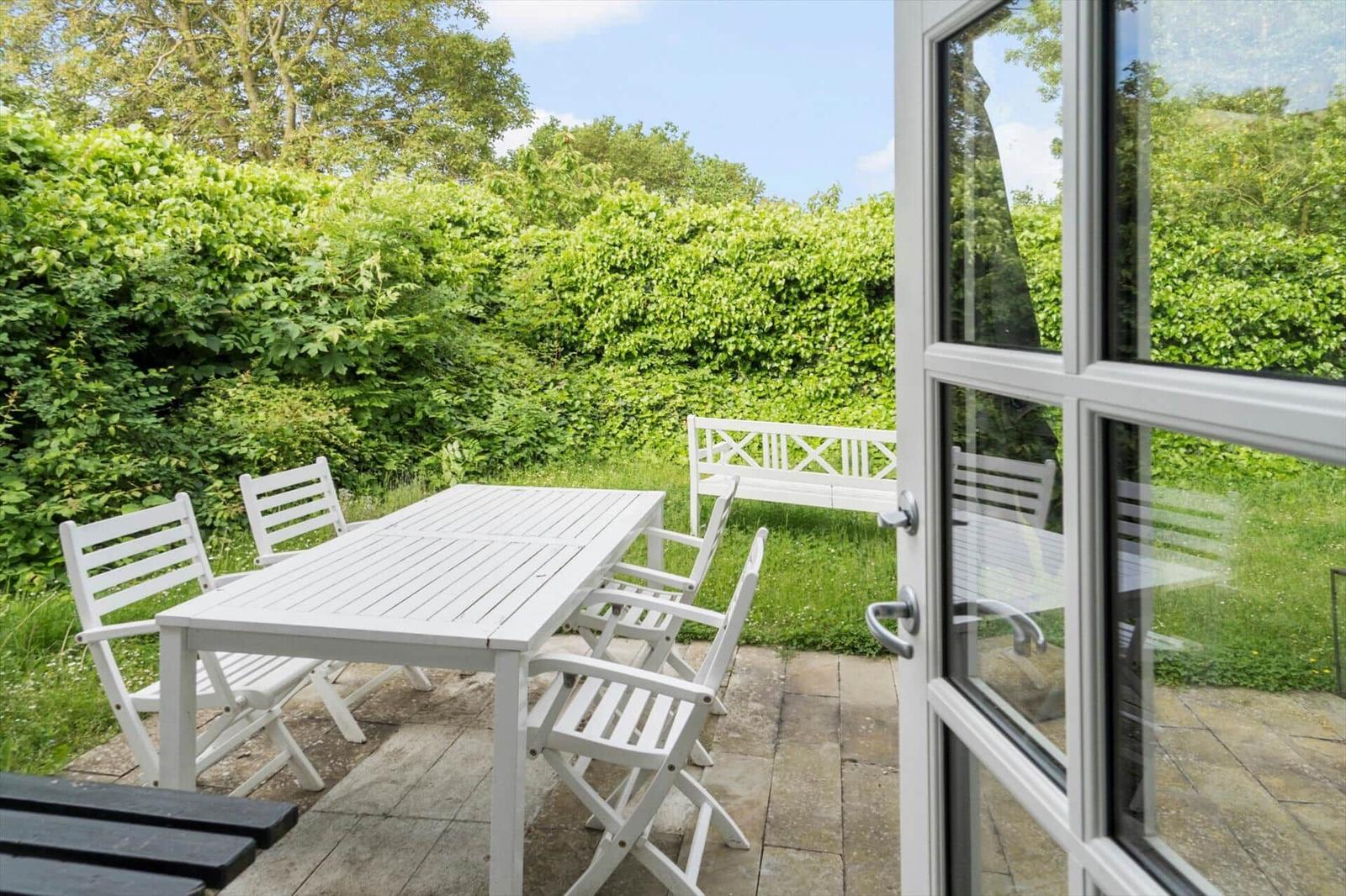 Terrasse mit weißem Tisch und Stühlen, umgeben von grüner Vegetation und einer Bank.