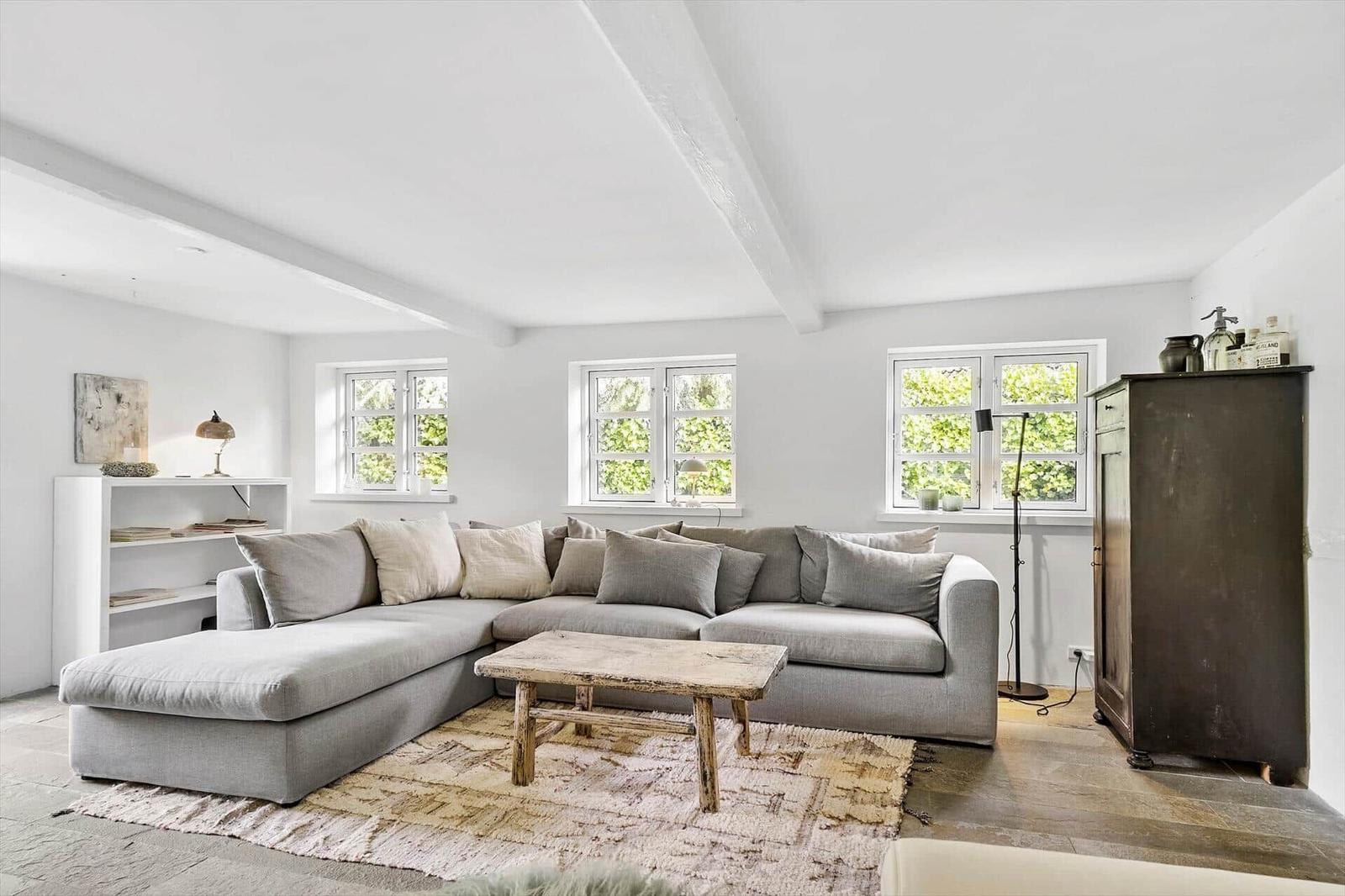 Wohnzimmer mit großem grauem Sofa, Holztisch und Fenstern mit Blick auf Grün.