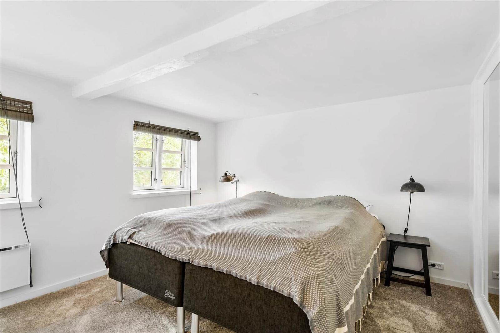 Schlafzimmer mit Bett, zwei Fenstern und Nachttisch mit Lampe.